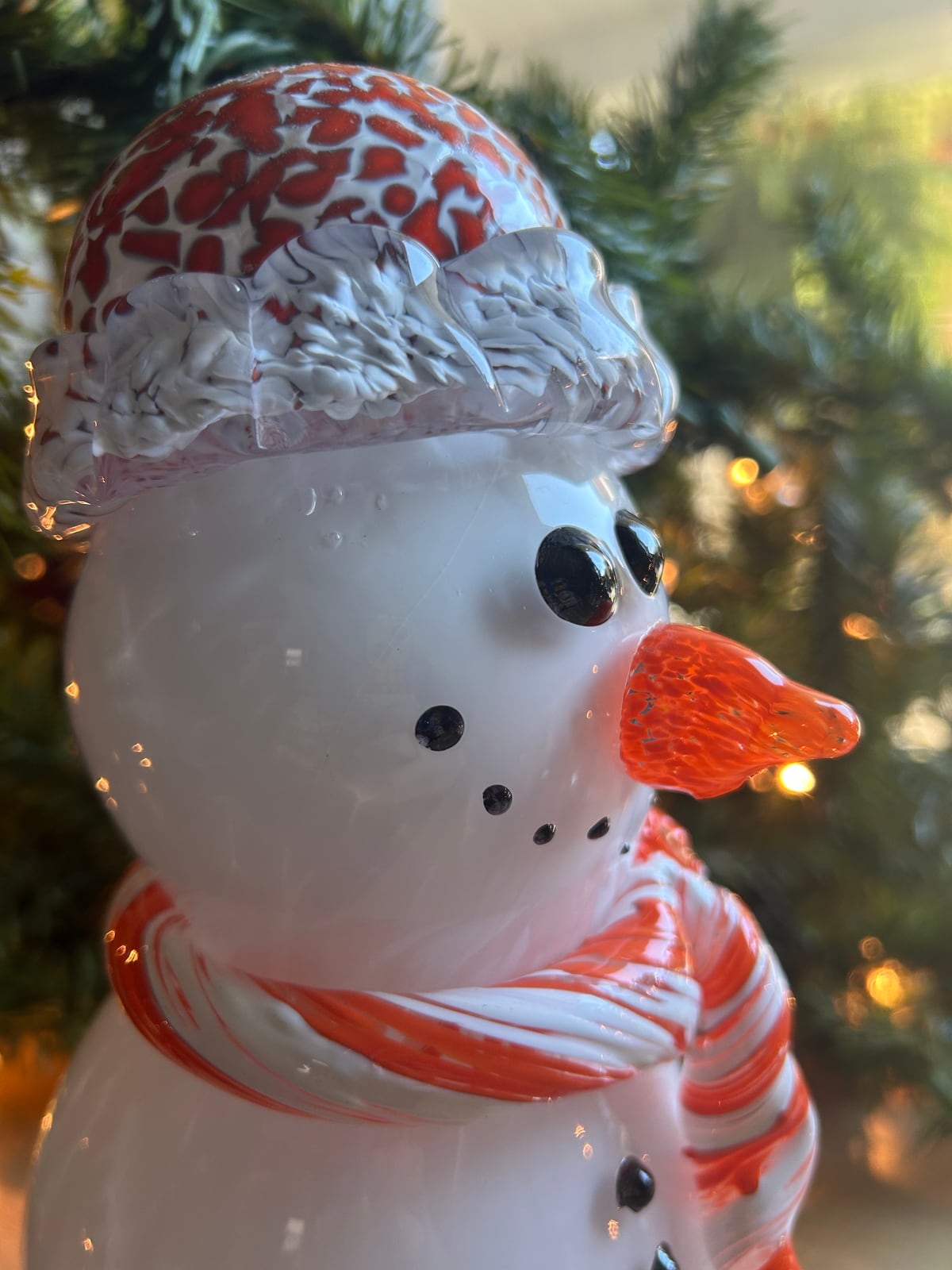 A glass snowman with a red patterned hat and red and white scarf beside tree lights
